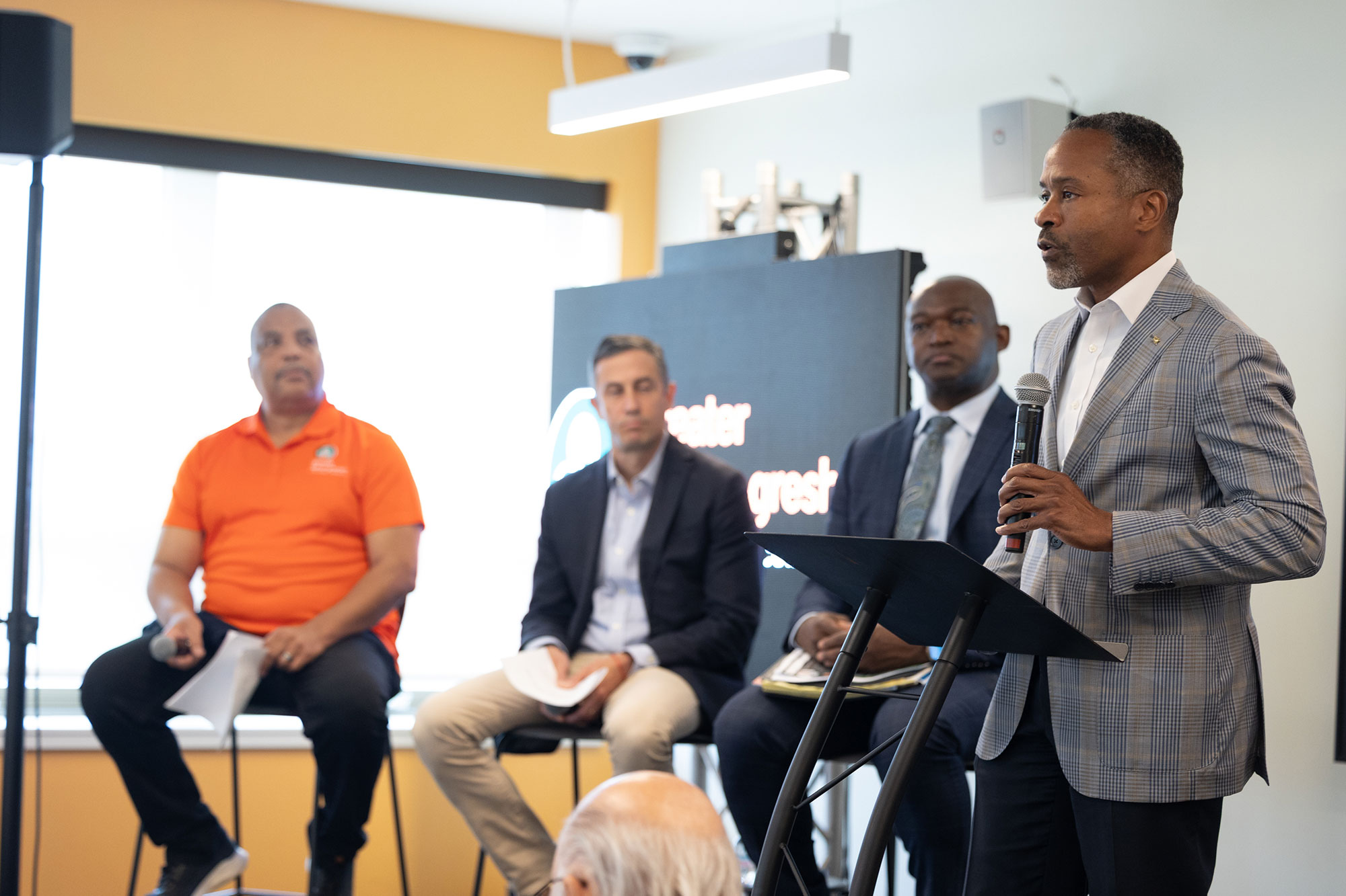 A person talks while standing on stage and three people sit next to them