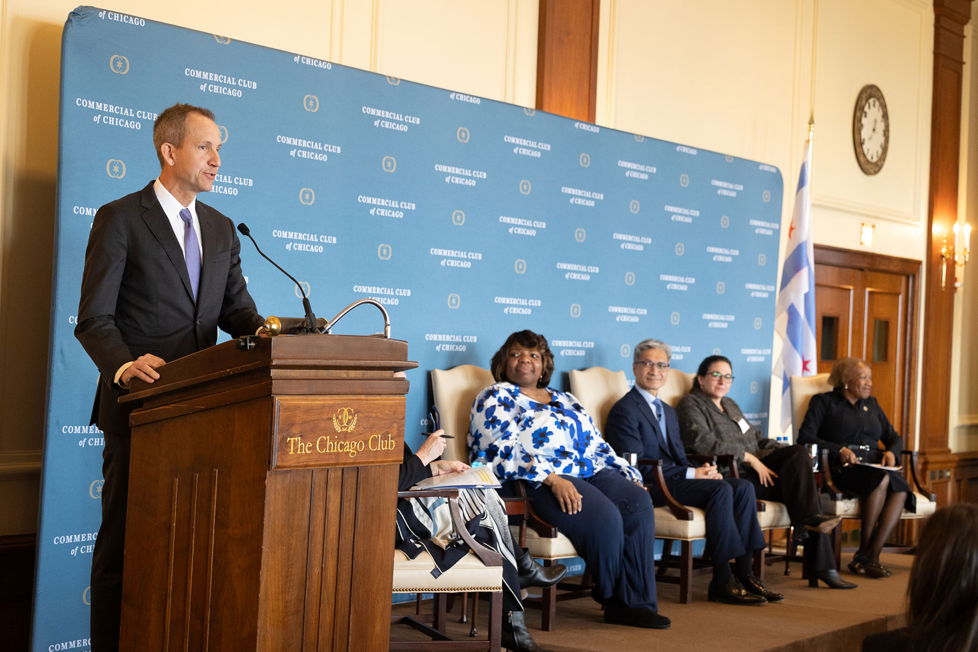 A person speaks at a podium with five people sitting in chairs on stage